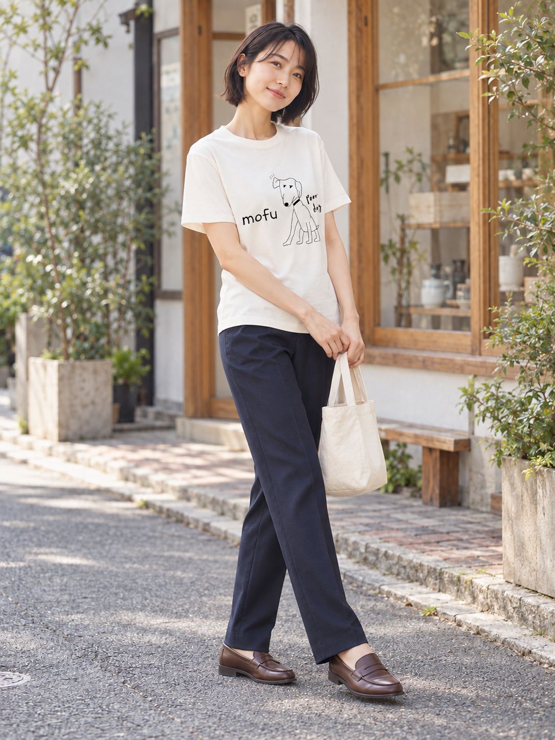布バッグを提げて、小さな店の前の道を歩く女性。白いTシャツに紺のパンツ